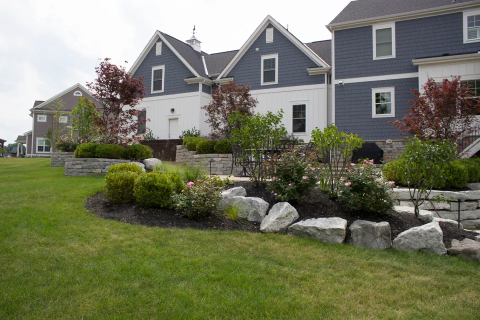 Low stone wall and boulders holding a planting bed above a lawn beside blue-gray houses